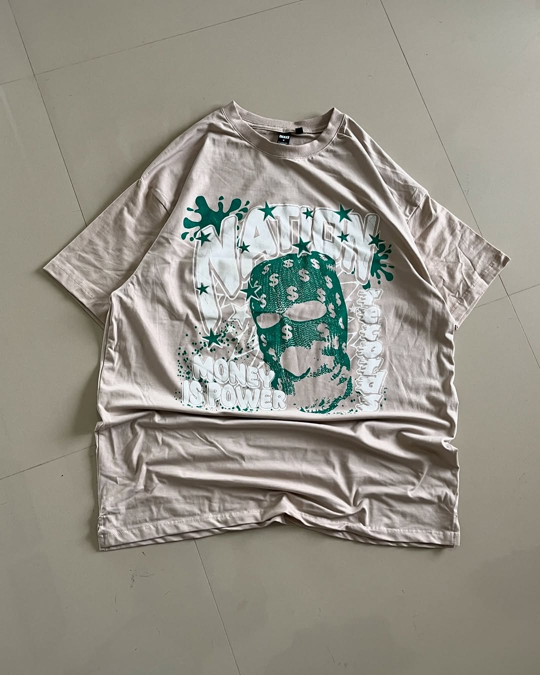 Peach oversized graphic tee featuring a teal balaclava mask with dollar signs and bold “NATION” and “MONEY IS POWER” text

