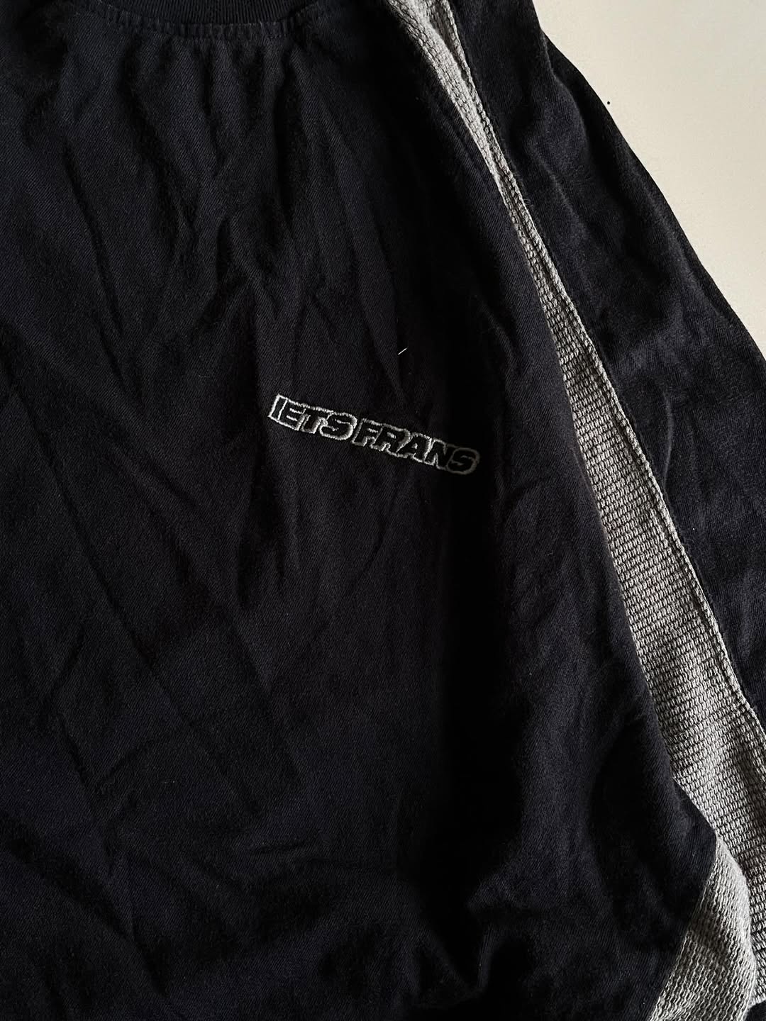 JUNKFITS black tee close-up showing embroidered 'IETS FRANS' logo and mesh texture.